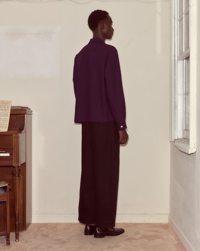 A person in dark clothing wears the Astor Trillium Shirt in purple, featuring men's wool fabric and frog closures, standing by a window and piano in neutral lighting, facing the wall.