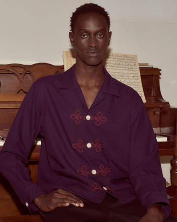 A person in an Astor Trillium Shirt - Purple sits in front of a piano with sheet music on the stand.