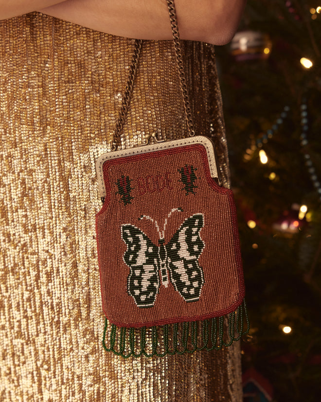 The Athalia Beaded Bag in Brown/Multi, adorned with intricate embroidered butterfly motifs and green fringe, hangs gracefully against a dazzling sequined background.