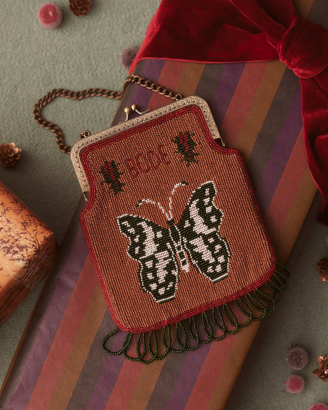 The Athalia Beaded Bag in Brown/Multi, adorned with intricate embroidered butterfly motifs and green fringe, hangs gracefully against a dazzling sequined background.