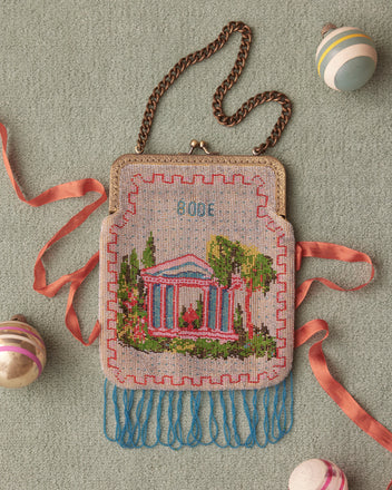 The Beaded Voyage Bag, adorned with a vintage sailboat design, features a metal chain strap and blue tassels, hanging elegantly against a tiled wall.