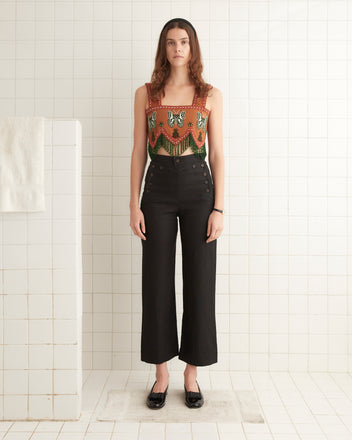 A person stands in a white-tiled room, wearing the Athalia Fringe Top and black high-waisted pants, with black shoes. The intricacy of the beadwork on this 100% cotton sleeveless top adds an elegant touch to their ensemble.