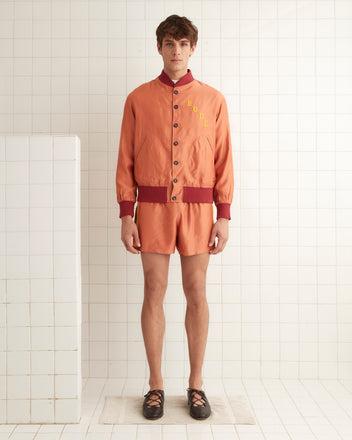 A person in a vibrant orange Rally Jacket and matching shorts stands against a pristine white tiled wall, bringing a burst of Manhattan style to the scene.