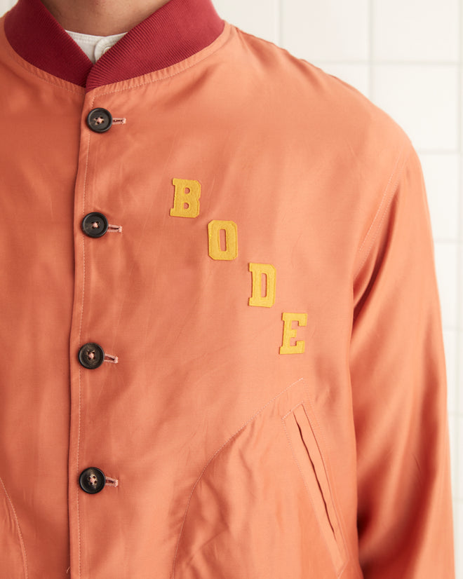 A person in a vibrant orange Rally Jacket and matching shorts stands against a pristine white tiled wall, bringing a burst of Manhattan style to the scene.