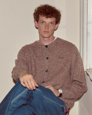 A young man with curly hair, wearing the Atlas Cardigan and jeans, sits indoors by a window, looking at the camera.