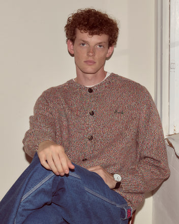 Young man with curly hair wearing the Atlas Cardigan and jeans, sitting indoors by a white wall and window.