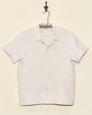 The Aubers Short Sleeve Shirt, a white button-up crafted from antique French linen, hangs on a hook against a plain light-colored wall.
