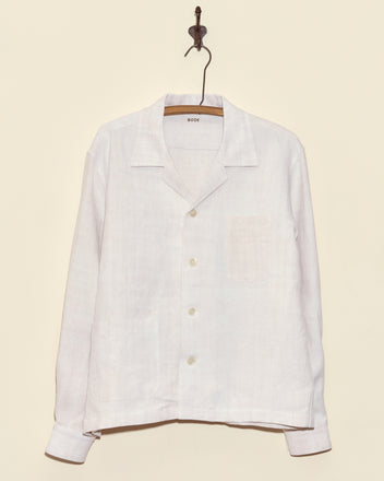 The Aubers Long Sleeve Shirt, crafted from antique French linen, hangs on a wooden hanger against a beige wall.