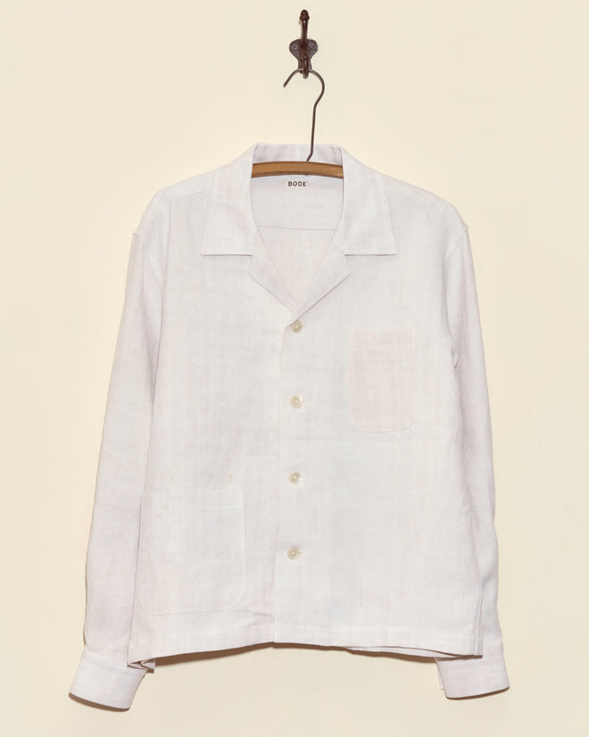 The Aubers Long Sleeve Shirt, crafted from antique French linen, hangs on a wooden hanger against a beige wall.