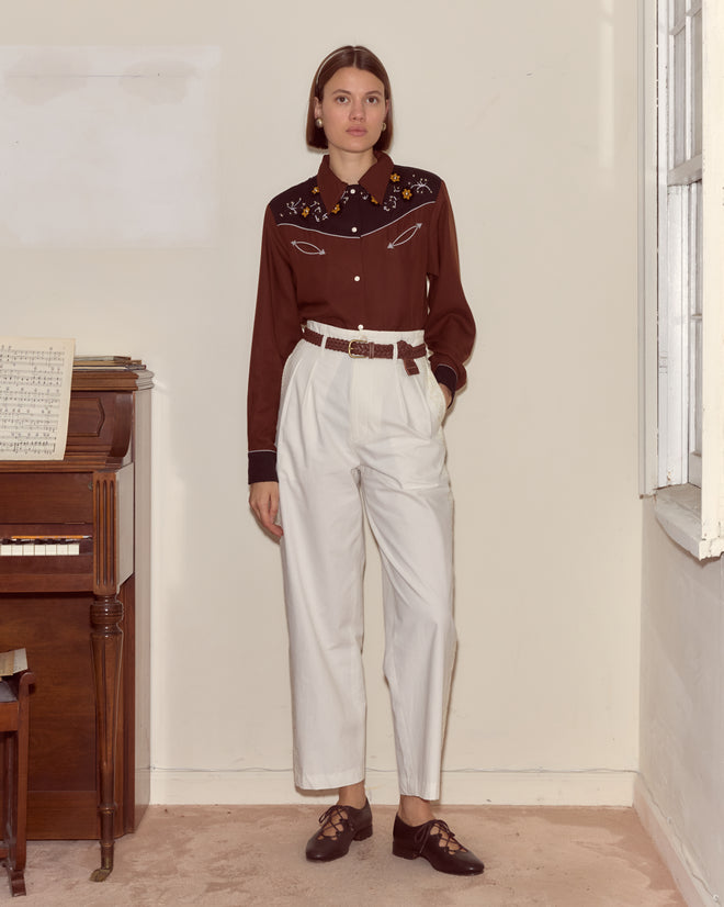 A woman in the Abilene Western Shirt and white trousers stands indoors by a piano and a window.