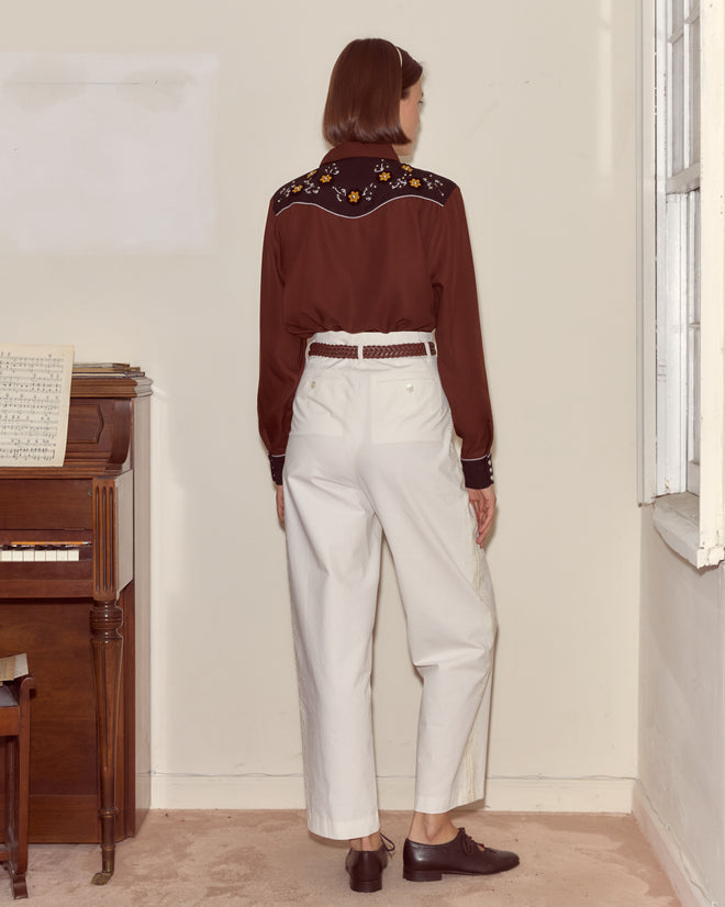 A woman stands indoors facing away, wearing the Abilene Western Shirt in brown with white pants near a piano and window.