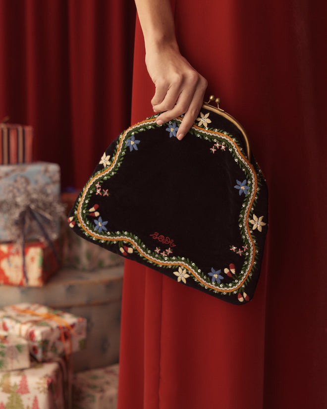 A person holds the Alpineflower Clutch against a red curtain, with wrapped gifts in the background, exuding elegance and sophistication.