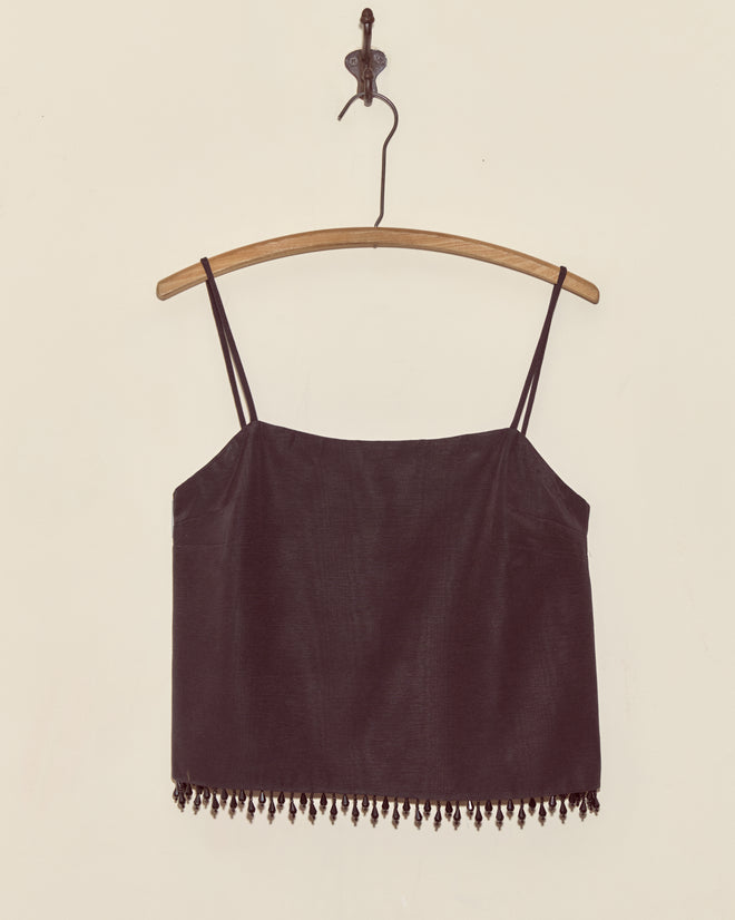 The Anglia Jet Tank - S is a black spaghetti strap crop top with droplet bead trim and a beaded fringe hem, evoking a vintage faille tank. Shown hanging on a wooden hanger against a plain wall.
