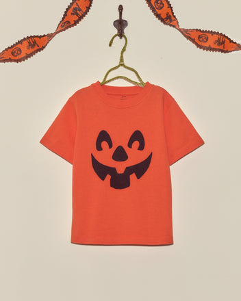 The Pumpkin Kids' Tee, featuring a bold black jack-o-lantern face, hangs on a hanger with Halloween garland above—making it the perfect festive vintage Halloween shirt for kids.