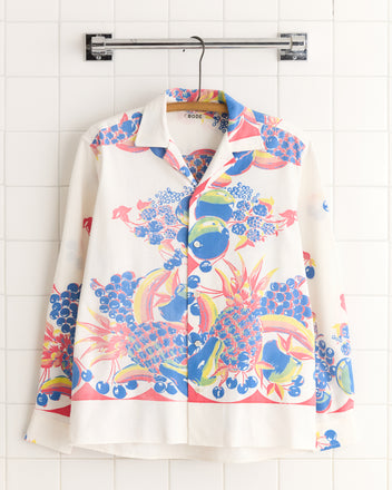 The Banana Bowl Shirt (XS/S) features a long-sleeve button-up design with a vibrant fruit and floral pattern, reminiscent of a vintage tablecloth, displayed on a hanger against white tiles—a unique statement piece.
