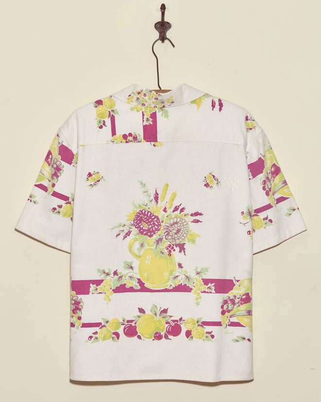 The Banana Bunch Short Sleeve Shirt (L/XL) features a white vintage tablecloth design with yellow and pink floral and fruit pitcher patterns and is shown hanging on a wall hook.