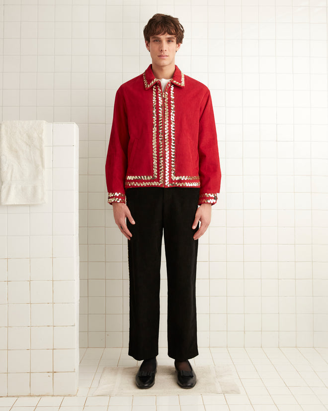 A person wearing a Bandstand Jacket in red, featuring gold sequin trim, stands in a tiled room next to a white towel on a rack.