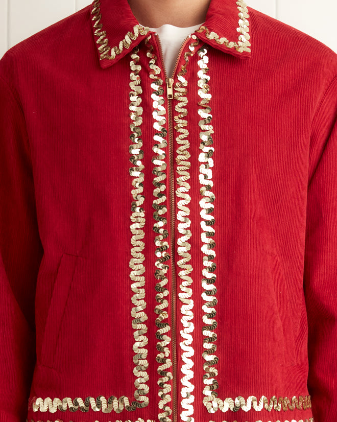 A person wearing a Bandstand Jacket in red, featuring gold sequin trim, stands in a tiled room next to a white towel on a rack.