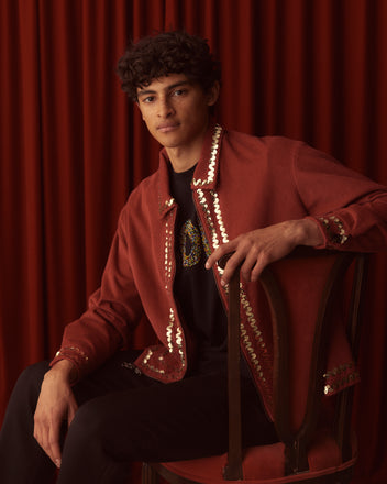 A person sits on a chair wearing the Bandstand Jacket - Red, featuring gold sequin trim and a classic workwear silhouette, in front of red curtains.