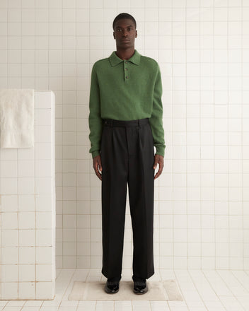 A man wearing a green sweater and black Barathea McNab Trousers, which evoke the wide-leg style of men's trousers from the 1940s-1950s, is standing in a tiled room with a white towel hanging on a nearby wall.