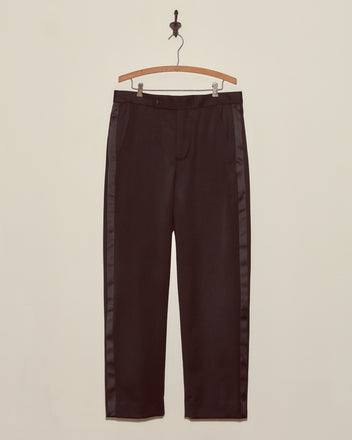 Barathea Tuxedo Trousers, crafted from 100% wool with a satin tuxedo stripe, hang on a wooden hanger against a plain beige wall.