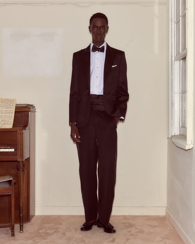 A man wearing the Barathea Single Breasted Tuxedo Jacket stands indoors by a piano and window on a beige carpet, embodying a stylish 1960s-inspired look.