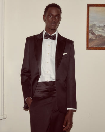 A person wearing the Barathea Single Breasted Tuxedo Jacket stands indoors by a framed painting, pairing the classic black jacket with a bow tie for a timeless 1960s-inspired look.