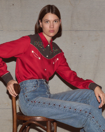 Woman wearing a red and black blouse with light wash jeans while sitting on a stool.