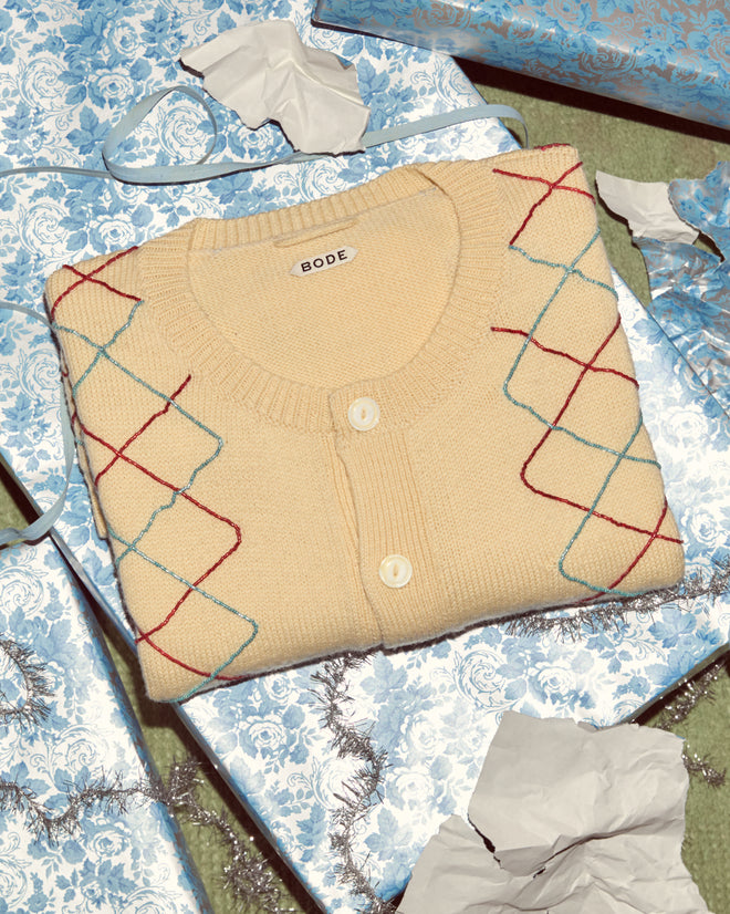 A folded Beaded Argyle Sketch Cardigan rests on blue gift-wrapped boxes, surrounded by torn wrapping paper.