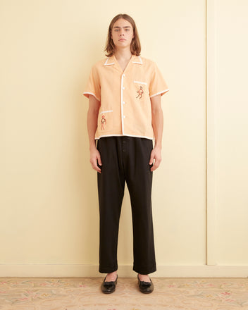 A person with long hair, dressed in a Beaded Blackjack Short Sleeve Shirt and black pants, stands against a light-colored wall on a patterned carpet.