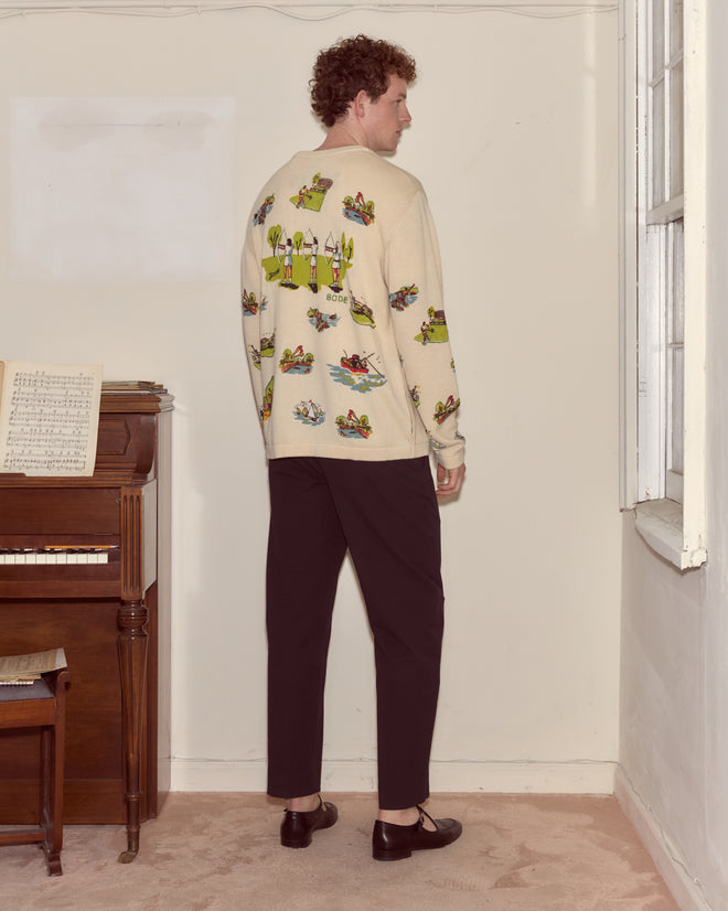 A man stands indoors by a piano, facing away, wearing the Beaded Camp Cardigan with frog illustrations and black pants.