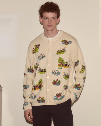 A man in a Beaded Camp Cardigan with colorful scenic prints stands against a plain beige wall.