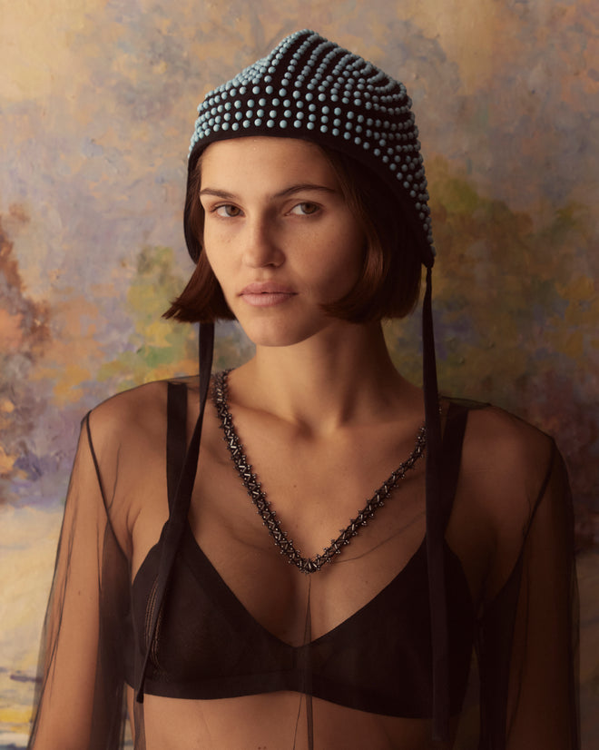 Wearing the Beaded Hat, a woman in a sheer top poses against an abstract, painted background.