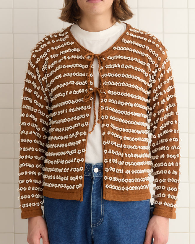 A person stands against a tiled wall wearing a Beaded Cardigan with 1950s-inspired brown and white stripes, adorned with floral patterns. Pearl-like beads highlight the hand-knit details, adding vintage elegance to the outfit.