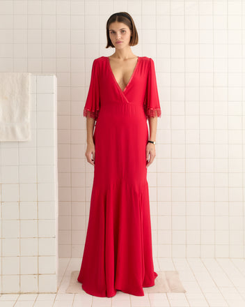 A woman in a 1930s-inspired Beaded Claret Gown - Red stands gracefully in a white-tiled room, with the textured crepe fabric and godet skirt enhancing elegance, while a towel hangs on the wall.