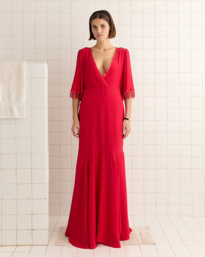 A woman in a 1930s-inspired Beaded Claret Gown - Red stands gracefully in a white-tiled room, with the textured crepe fabric and godet skirt enhancing elegance, while a towel hangs on the wall.