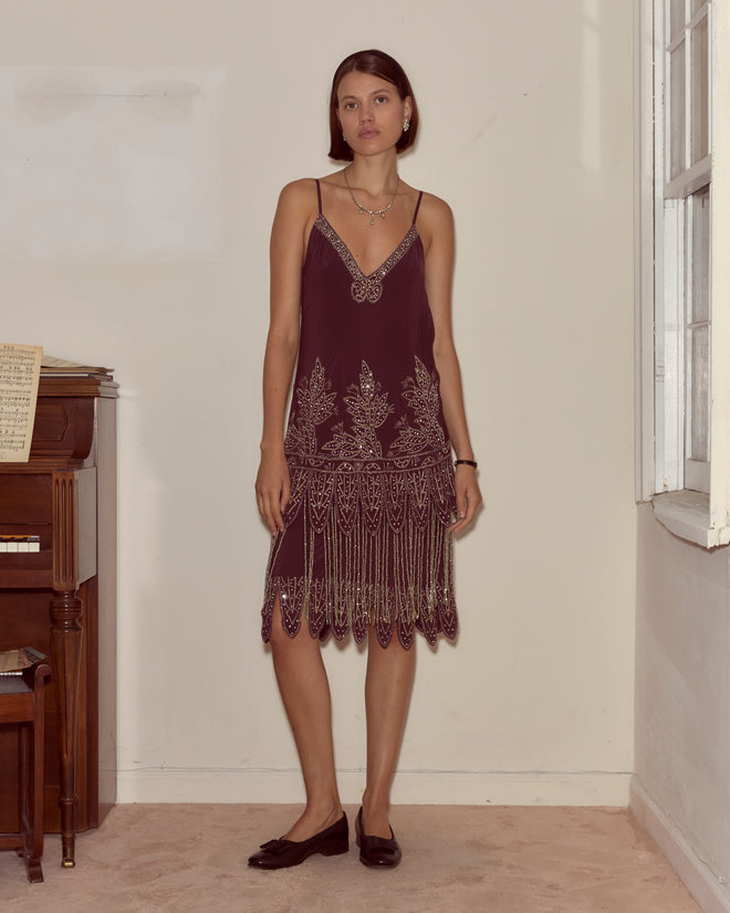 A woman in a Beaded Epoque Dress stands indoors by a piano and window, looking at the camera.