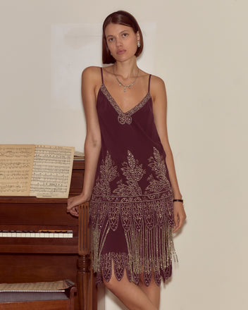 A woman in the Beaded Epoque Dress, featuring burgundy silk, beading, and gold embroidery, leans on a piano with sheet music against a plain wall.