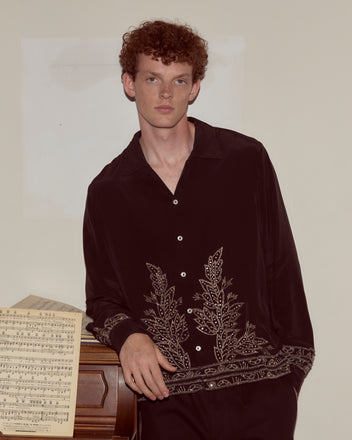 A young man wears the Beaded Epoque Long Sleeve Shirt as he leans on a piano with sheet music, set against a plain wall.