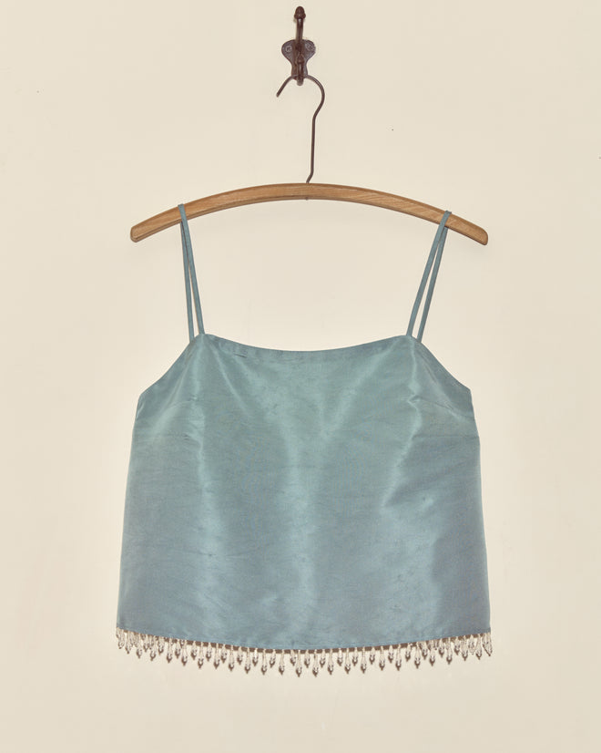The Beaded Frost Tank - S is a light blue satin camisole with thin straps and droplet beads, reminiscent of a vintage faille tank, displayed on a wooden hanger against a plain wall.