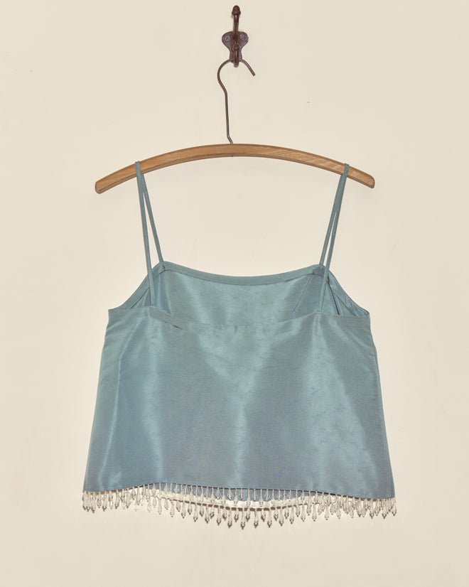 The Beaded Frost Tank - S is a light blue satin camisole with thin straps and droplet bead fringe, displayed on a wooden hanger against a wall.