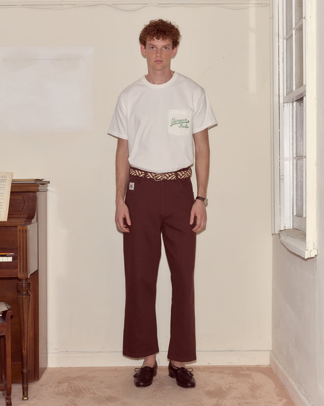 A man wears the Beaded Geneva Pocket Tee, a 100% cotton white T-shirt, and brown pants as he stands indoors near a piano and window, looking at the camera.