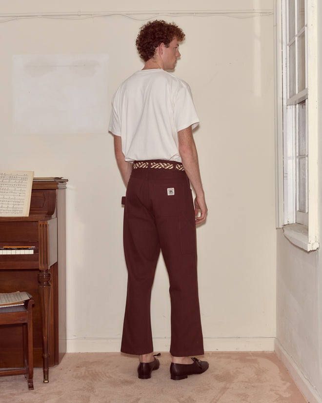 A man faces away in a room near a piano and window, wearing the Beaded Geneva Pocket Tee in white and dark pants.