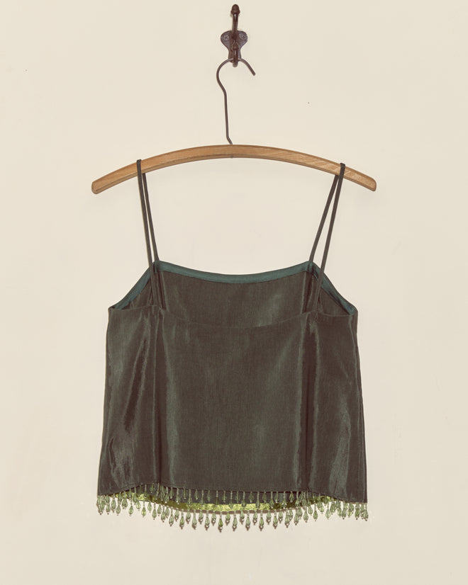 The Beaded Hemlock Tank - S is a dark green velvet camisole with thin straps and droplet bead fringe, displayed on a wooden hanger against a beige wall. Made in New York for timeless sophistication.