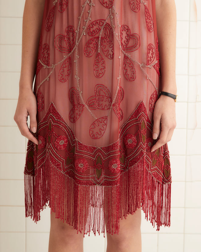 In a tiled room, a person faces the camera wearing the Beaded Ilex Dress—an intricately designed silk dress in red with fringe detailing and exquisite hand beadwork.