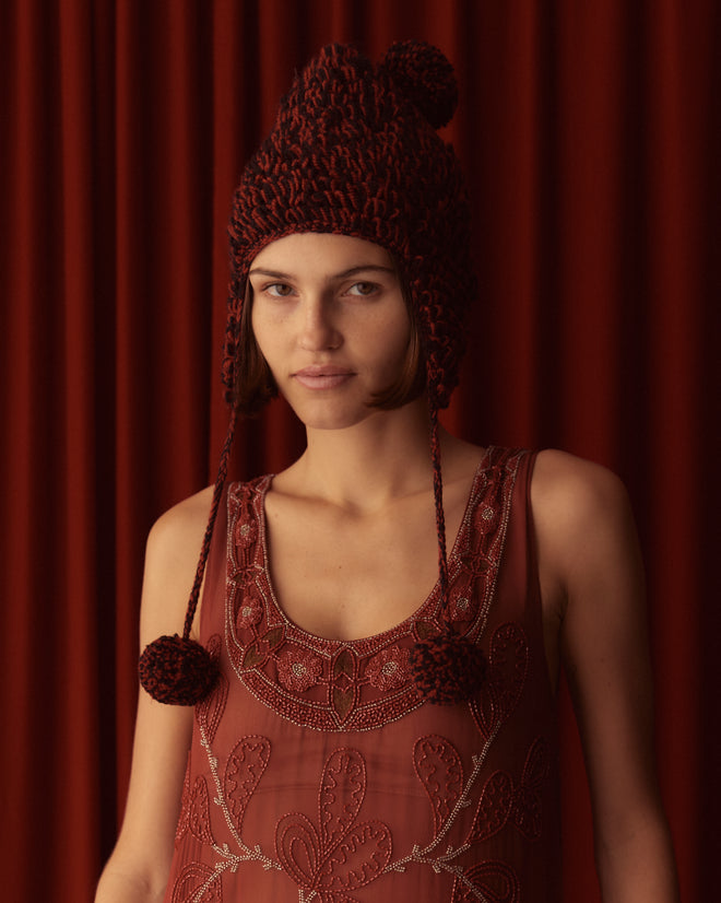 A person wearing the Loopy Tassel Hat featuring pom-pom tassels and an embroidered top stands in front of a matching red curtain.