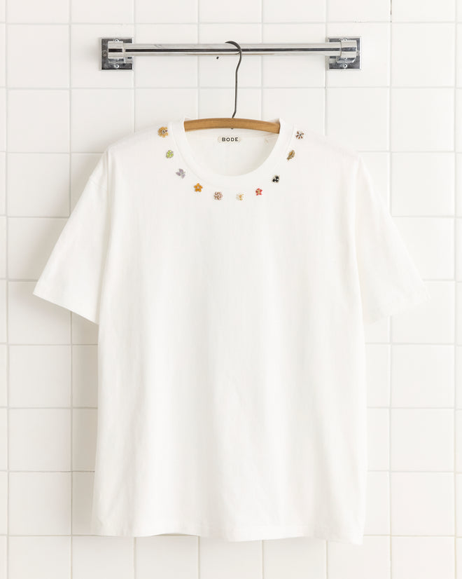 The Beaded Necklace Tee is a white 100% cotton t-shirt featuring small colorful flower embroidery and unique vintage sari-inspired appliqué details around the neckline, displayed on a wooden hanger.