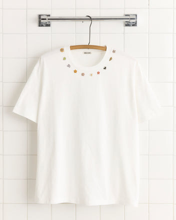 The Beaded Necklace Tee is a white 100% cotton T-shirt with small, colorful embroidered flowers around the neckline, shown hanging on a hanger.