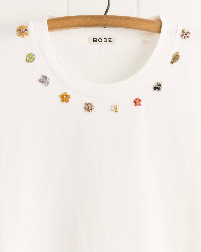 The Beaded Necklace Tee by BODE, made from 100% white cotton, features small colorful beaded floral pins adorning the neckline for a unique textile touch and hangs on a wooden hanger.