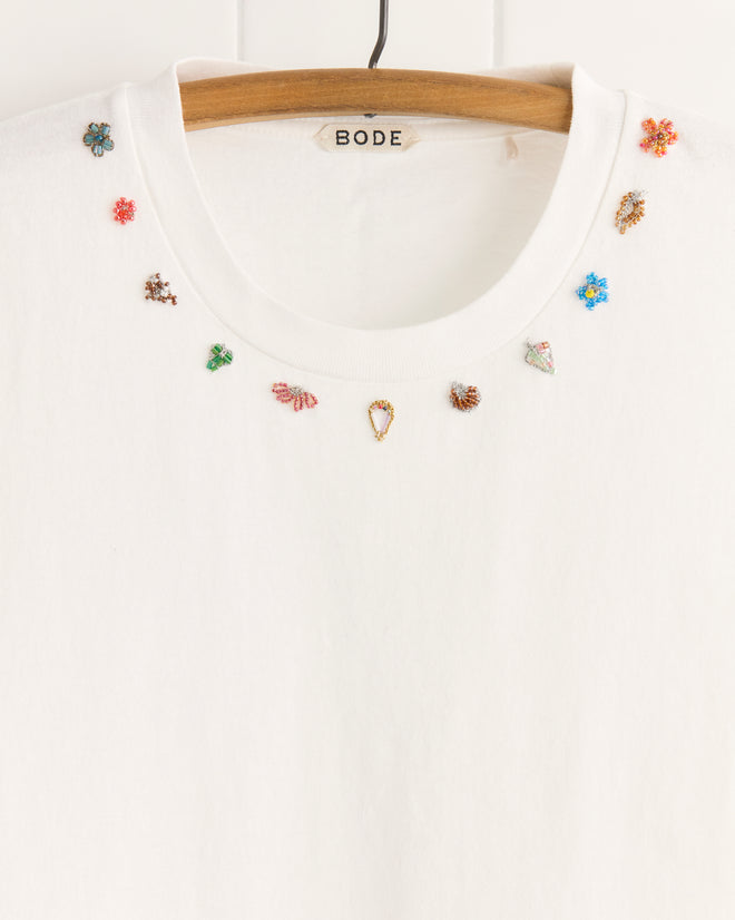 The Beaded Necklace Tee is a white, 100% cotton t-shirt with small, colorful beaded insect and flower designs around the neckline, inspired by unique textile details and displayed on a wooden hanger.